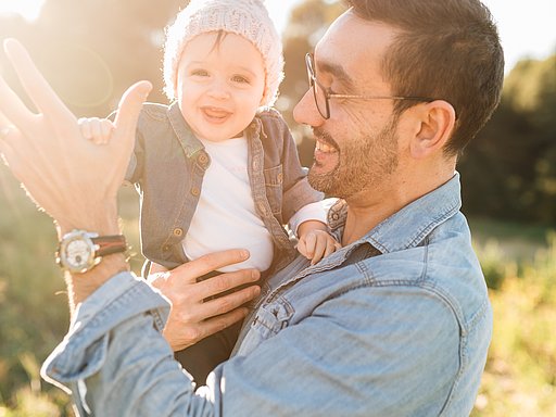 Ein Mann steht auf einem Feld und hält lachend sein Kleinkind auf dem Arm.